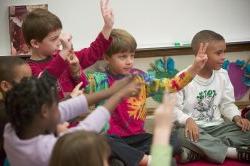 Image of children sitting on the floor in a classroom, raising their arms.